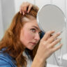 Woman examining thinning hairline in mirror before hair restoration in Saraland AL