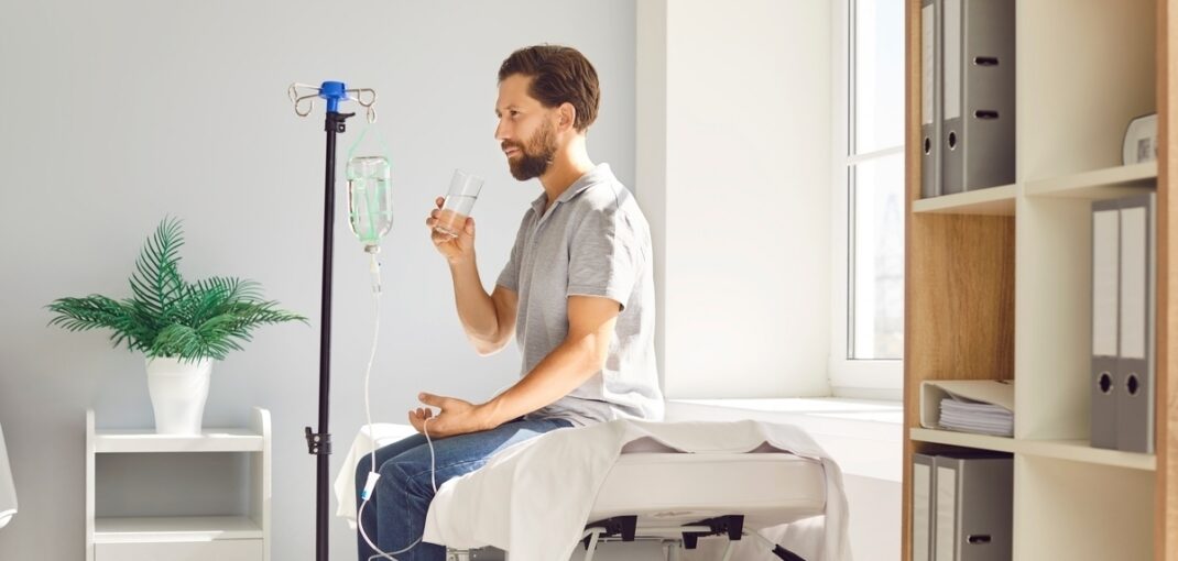 Man holding a glass of water while connected to an IV drip representing IV hydration vs drinking water comparison