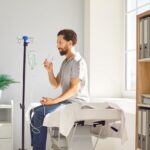 Man holding a glass of water while connected to an IV drip representing IV hydration vs drinking water comparison