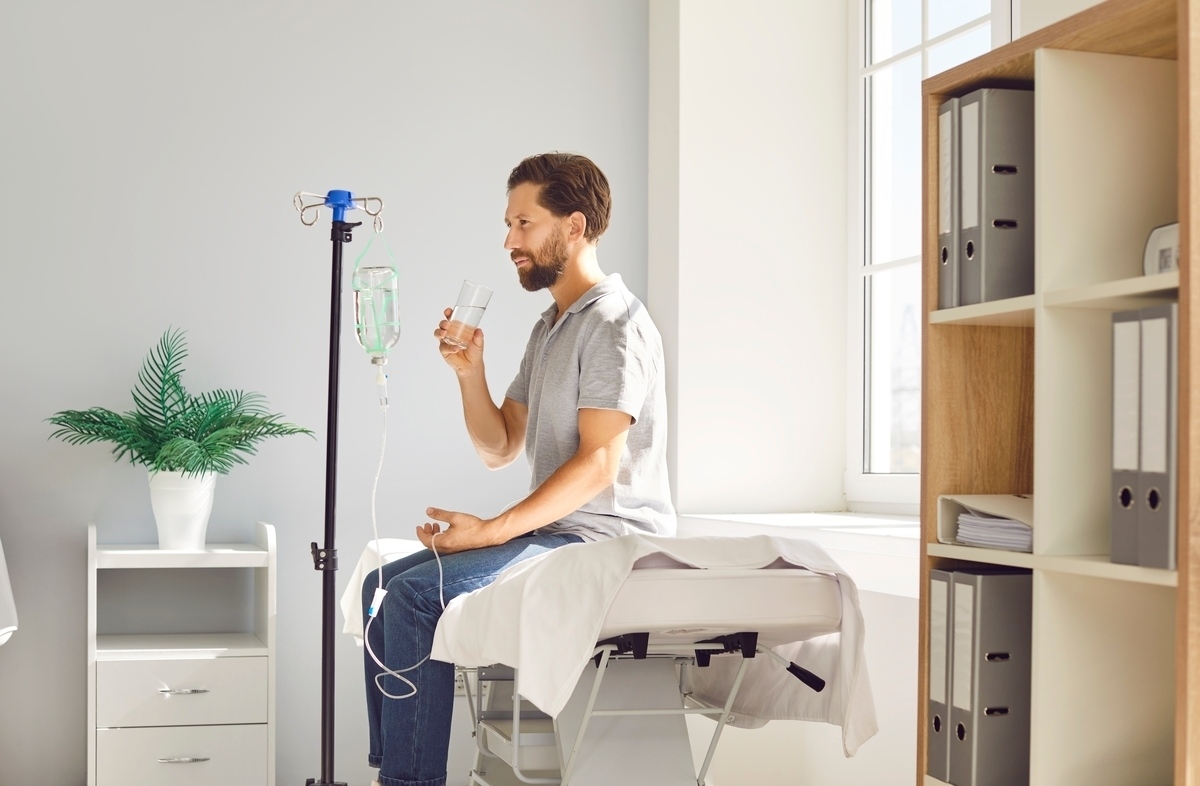 Man holding a glass of water while connected to an IV drip representing IV hydration vs drinking water comparison