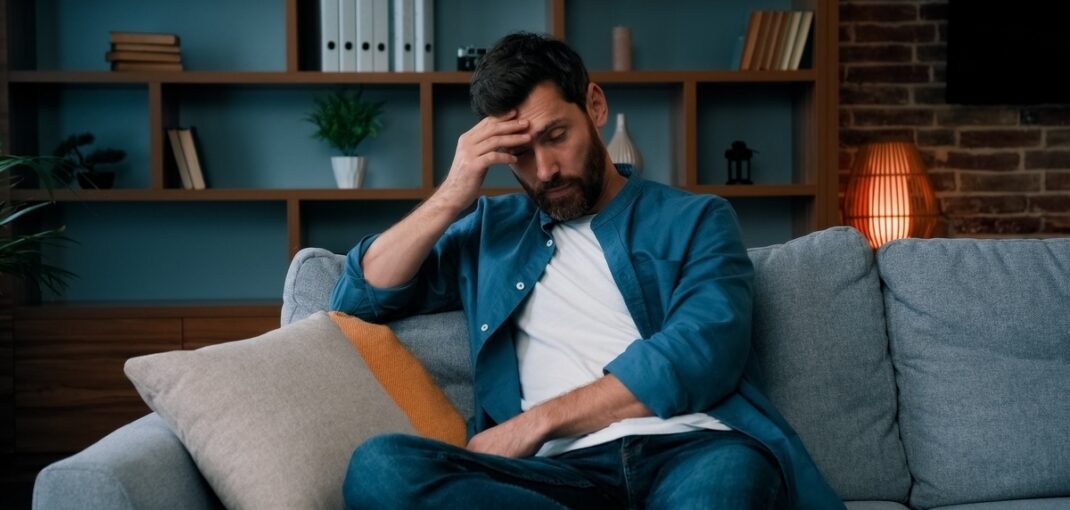 Tired bearded man sitting on couch with hand on forehead showing symptoms that peptide therapy for men can help address
