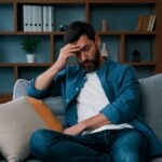 Tired bearded man sitting on couch with hand on forehead showing symptoms that peptide therapy for men can help address