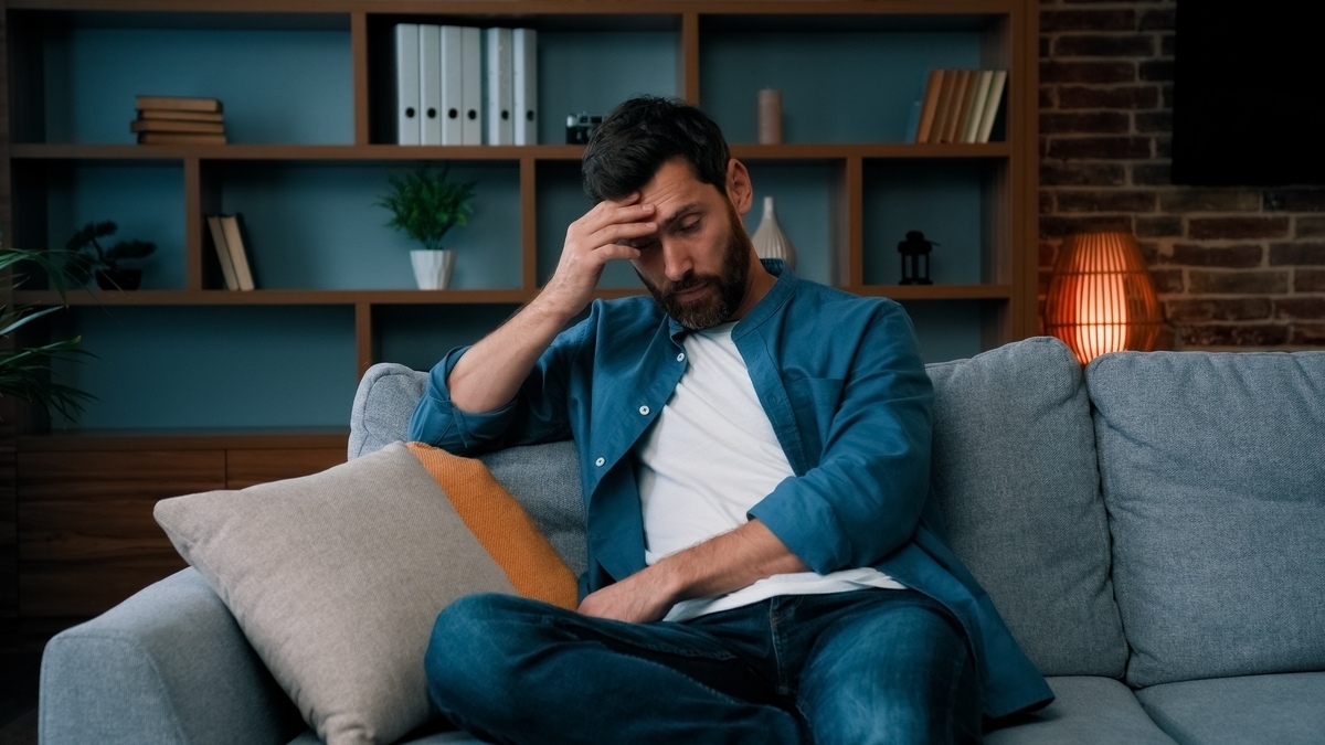 Tired bearded man sitting on couch with hand on forehead showing symptoms that peptide therapy for men can help address